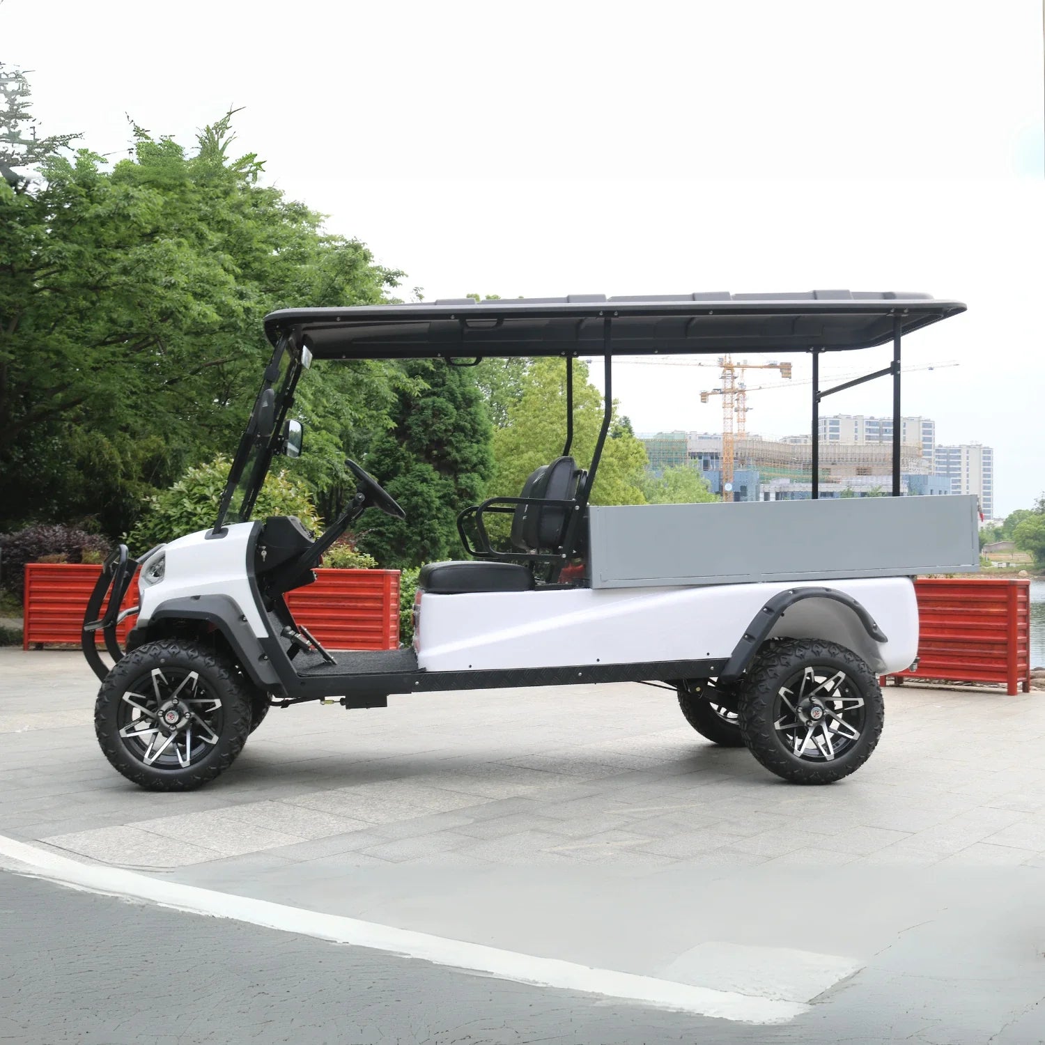 ocean cartz full side view of a long-bed white utility golf cart with a black roof and a large, grey rear cargo box. This six-passenger electric vehicle is a lifted off-road cart featuring black seats, a front brush guard, and aggressive all-terrain tires. The cart is equipped with custom black and silver wheels and is parked outdoors with trees and urban construction visible in the background.