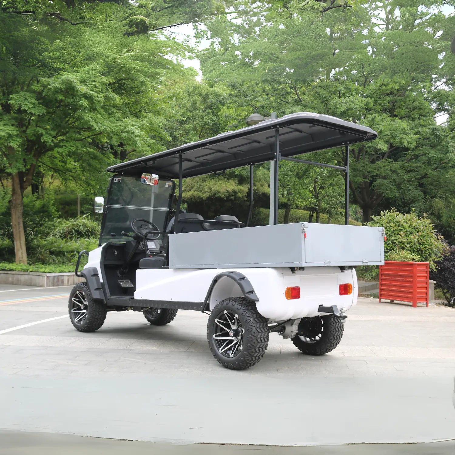 ocean cartz rear three-quarter view of a white utility golf cart with a black roof and a large, grey rear cargo box. This lifted electric vehicle is an off-road work cart featuring aggressive all-terrain tires on custom black and silver wheels. The two-seater cart is shown on a paved road surrounded by lush green trees and foliage in a park or large campus environment.