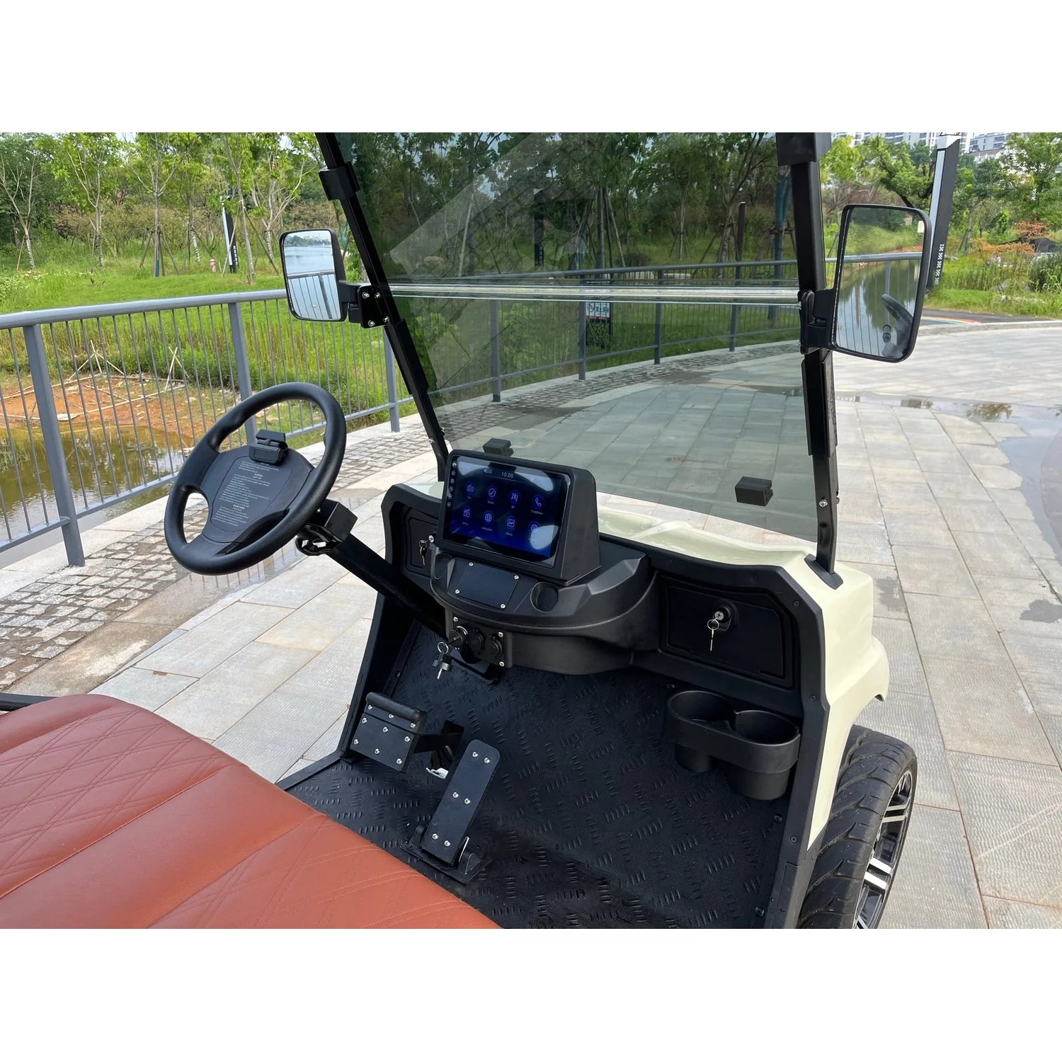 ocean cartz close-up interior view of a cream-colored luxury golf cart cockpit, focusing on the steering wheel and dashboard. The image highlights the digital touchscreen display mounted centrally, a black steering wheel, two cup holders, and textured black floor mats. The driver's area shows a brown seat edge. The electric vehicle is parked outdoors on a paved area overlooking a pond or waterway, emphasizing its features as a premium NEV or resort transport cart.