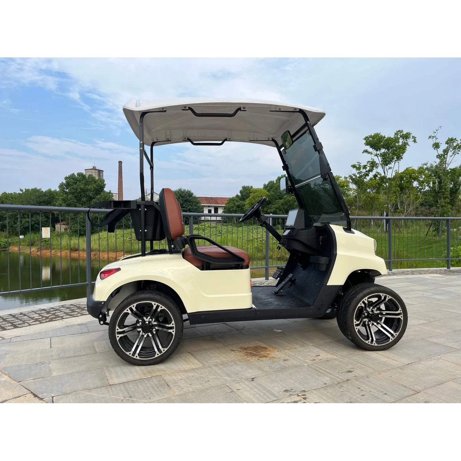 ocean cartz side profile view of a cream-colored four-seater luxury golf cart with a white roof, parked on a paved stone path next to a dark body of water. This electric vehicle features brown and black seats, a modern front end, and a tinted windshield. The lifted golf cart is equipped with low-profile tires and custom black and silver alloy wheels, set against a partially industrial background with trees and a metal railing, highlighting its stylish NEV or resort transport design.