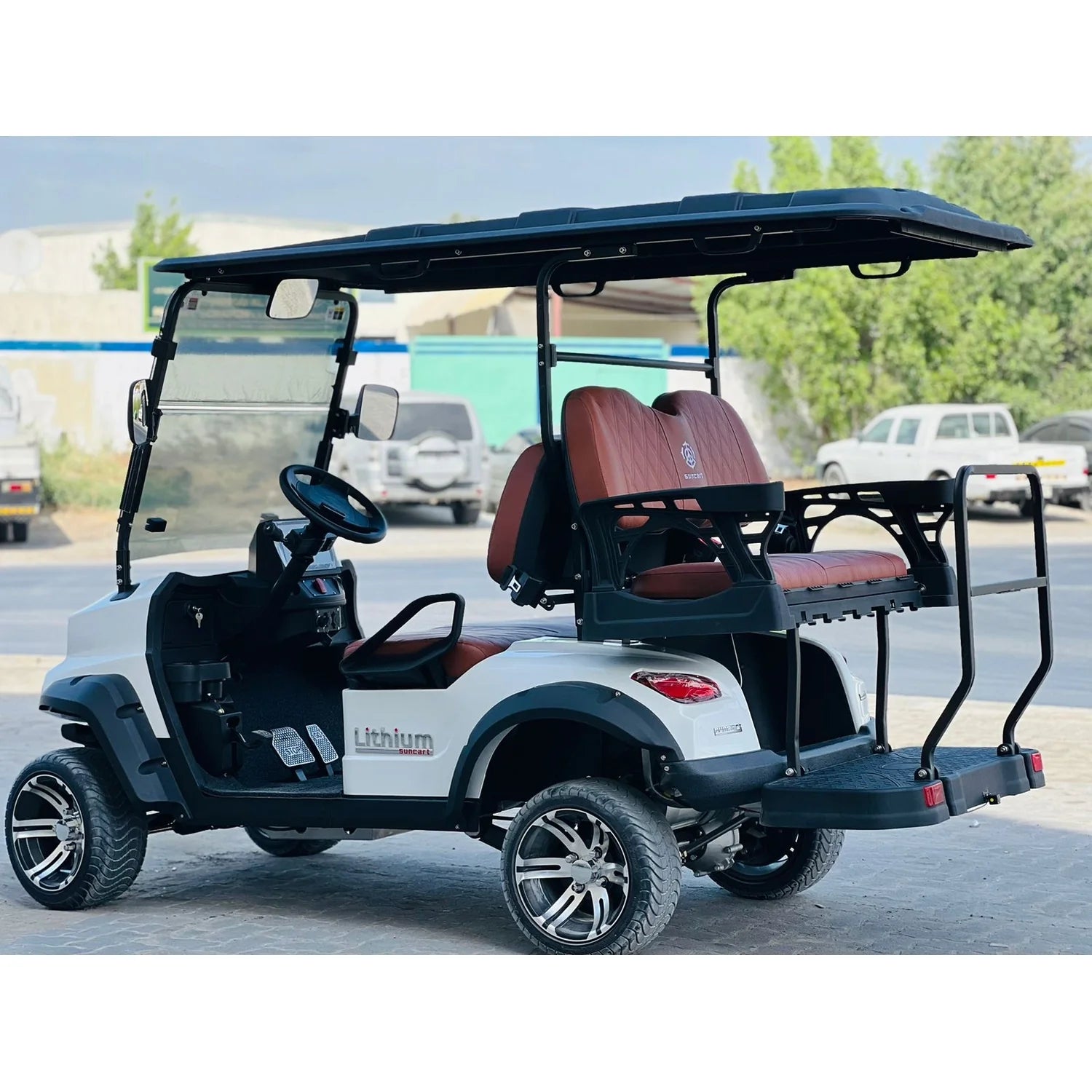 The cart has brown, diamond-stitched seats and a rear cargo area or seat with a black safety bar and grab handles. The side of the cart has a "Lithium Extreme" badge. It is fitted with custom silver and black alloy wheels and thick, all-terrain tires. The background is an outdoor industrial or storage area with a light-colored building, some utility vehicles, and trees under an overcast or hazy sky.