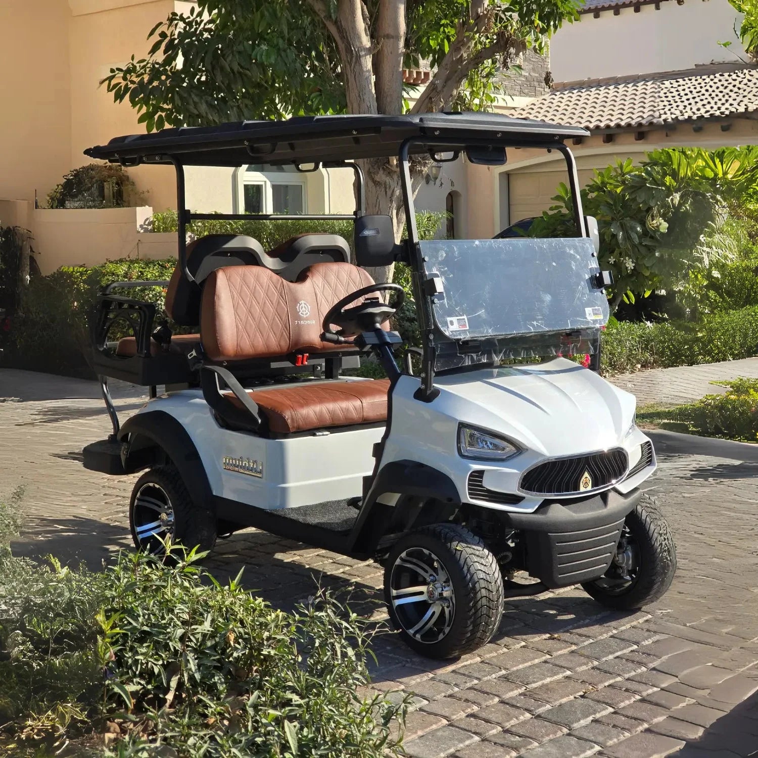 The cart has brown, diamond-stitched leather seats and a black roof. It features a modern, automotive-style front end with distinct headlights and a black grille. The cart is equipped with off-road tires and custom black and silver alloy wheels. Green bushes and plants frame the foreground, and a light-colored stucco house is visible in the background. The overall scene suggests a luxurious community or resort environment.