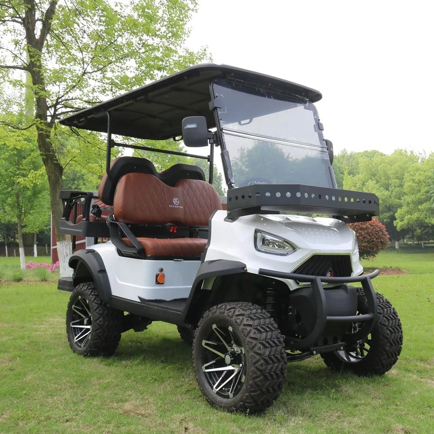 ocean cartz three-quarter front view of a white six-seater golf cart with a black roof and black trim, parked on a grassy area. This lifted electric vehicle is an off-road luxury golf car featuring brown diamond-stitched leather seats, a front brush guard, and an aggressive front design. It is equipped with large, knobby all-terrain tires on custom black and chrome wheels.