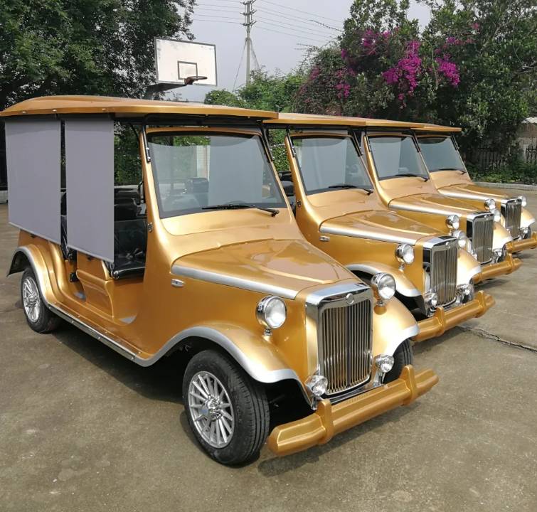 oceancartz three-quarter front view of a line of three gold classic antique-style golf carts with black roofs, parked on a paved court. The front vintage electric vehicle features a gold grille and bumper, silver wheels, and a pulled-down grey side panel for shade. These multi-passenger classic carts are shown in an outdoor setting with a basketball hoop and trees in the background, highlighting their use as resort transport vehicles.