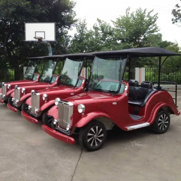 oceancartz three-quarter front view of a line of three dark red classic antique-style golf carts with black roofs, parked on a paved court. The front vintage electric vehicle features a large chrome grille, round headlights, black seats, and custom black and white wheels. These multi-passenger classic carts are shown in an outdoor park setting with a basketball hoop and trees in the background, highlighting their use as resort transport vehicles.
