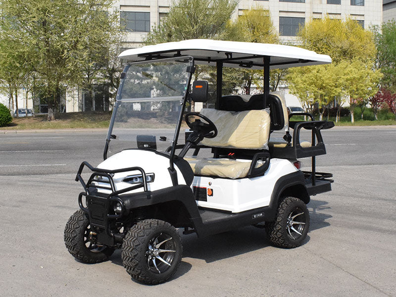 ocean cartz three-quarter front view of a white six-seater golf cart with a white roof, parked on an asphalt street. This lifted off-road electric vehicle features a black front brush guard, tan or cream-colored seats, and a rear flip seat. The all-terrain golf cart is equipped with large, knobby off-road tires on custom black and silver wheels, set against a background of trees and modern buildings.