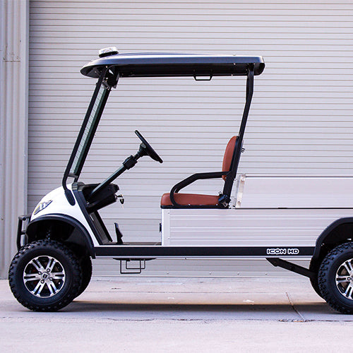 oceancartz side profile view of a white utility electric vehicle with a black roof and a silver aluminum rear cargo bed, parked in front of a light gray roll-up garage door. This specialized two-seater work cart features brown seats and a lifted suspension. The electric utility golf cart is equipped with all-terrain tires and custom black and silver wheels, highlighting its heavy-duty property maintenance function.