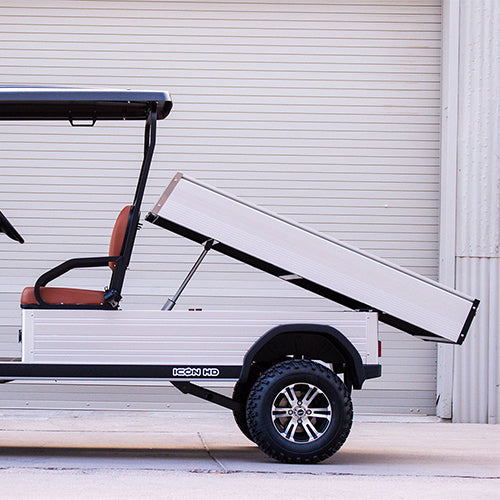 oceancartz side profile close-up of a white utility electric vehicle demonstrating its fully raised dump bed function, parked in front of an industrial roll-up door. This specialized two-seater electric work cart features brown seats and a silver aluminum rear cargo bed with a hydraulic lifting mechanism clearly visible. The lifted utility golf cart is equipped with all-terrain tires on custom black and silver wheels, highlighting its heavy-duty hauling and dumping capability.