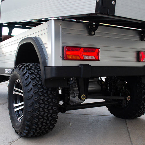 oceancartz close-up rear view of the silver aluminum cargo bed and the off-road tires on a lifted electric utility vehicle. This shot highlights the robust rear suspension, the black and silver custom wheel, and the large, knobby all-terrain tire. The rear panel of the cargo bed features a modern, rectangular LED taillight, emphasizing the heavy-duty and rugged design of the electric work cart.