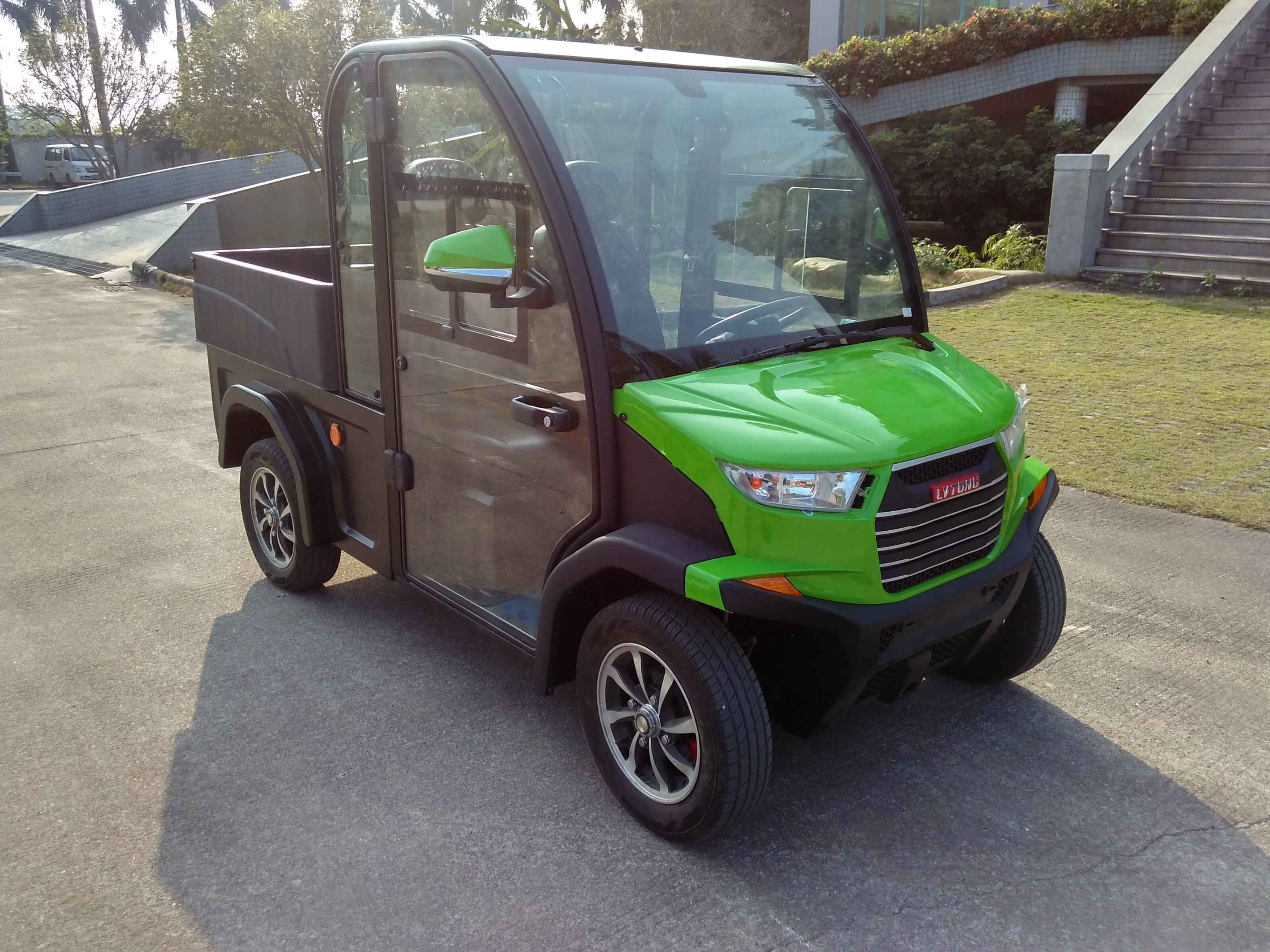 oceancartz three-quarter front view of a lime green and black utility electric vehicle with a fully enclosed cab and a black rear cargo bed. This two-seater commercial NEV or electric work cart features a bright green hood, black enclosed doors with glass, and a large front windshield. It is equipped with custom black and silver wheels and is parked on an asphalt and concrete area outside a modern building with steps and landscaping, highlighting its use as a compact utility transport vehicle.