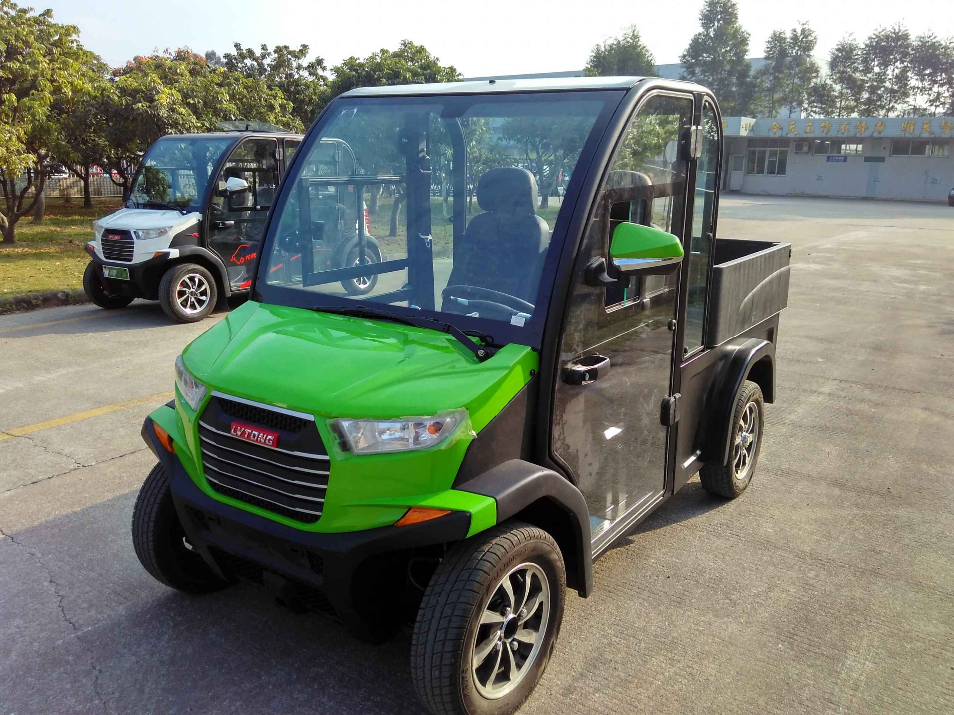 oceancartz three-quarter front view of a lime green and black utility electric vehicle with a fully enclosed cab and a black rear cargo bed, parked on an asphalt lot. This two-seater commercial NEV or electric work cart features a bright green hood, black enclosed doors with glass, and a modern front grille. Another similar vehicle is parked in the background, suggesting a fleet. The utility transport vehicle is outdoors with trees and buildings in the distance.