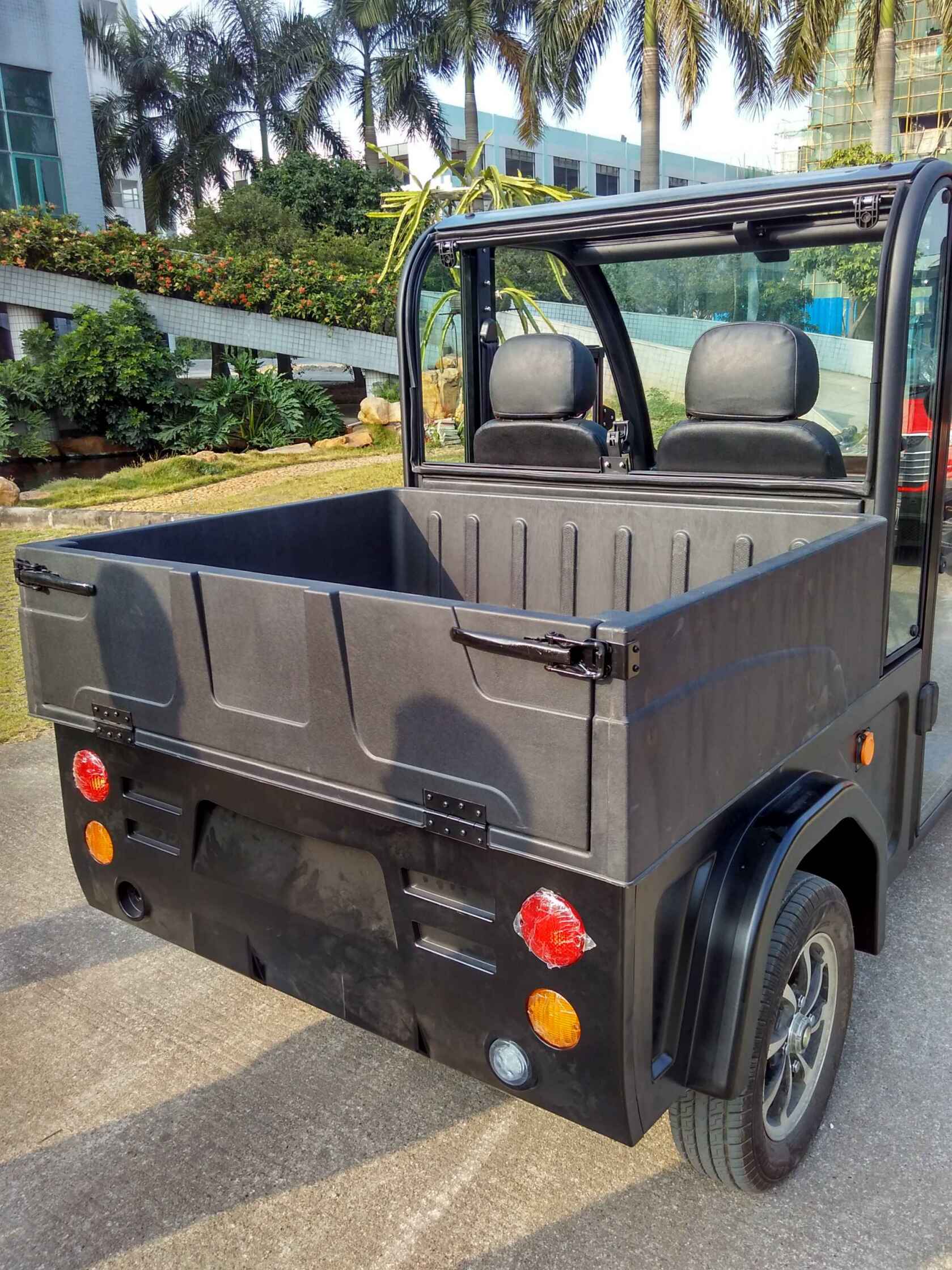 oceancartz rear view of a black electric utility vehicle with a durable, molded plastic cargo box and an open-top cabin, parked outdoors. This two-seater electric work cart features black seats, a clear rear window, and round red and amber taillights. The image highlights the deep hauling capacity of the utility transport vehicle, set in a sunny area with palm trees and a modern building in the background.